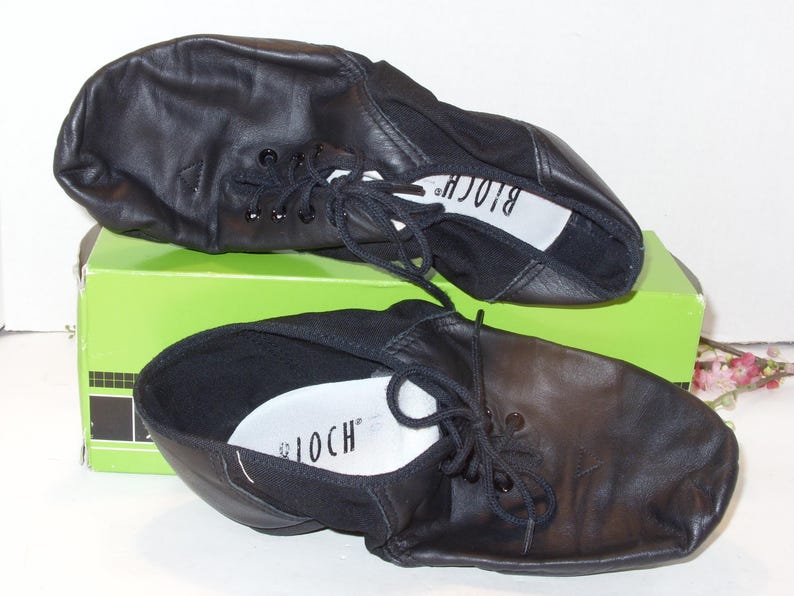 Puede incluir: Un par de zapatos de baile de cuero negro con cordones, apoyados en una caja verde. Los zapatos tienen una puntera redondeada y una suela blanda, con la marca "BLOCH" visible en la plantilla. Zapatos de baile.