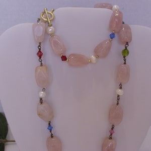 May include: A long necklace featuring large, light pink rose quartz beads, interspersed with small white pearls and colorful crystal accents. The necklace has a gold-tone toggle clasp.