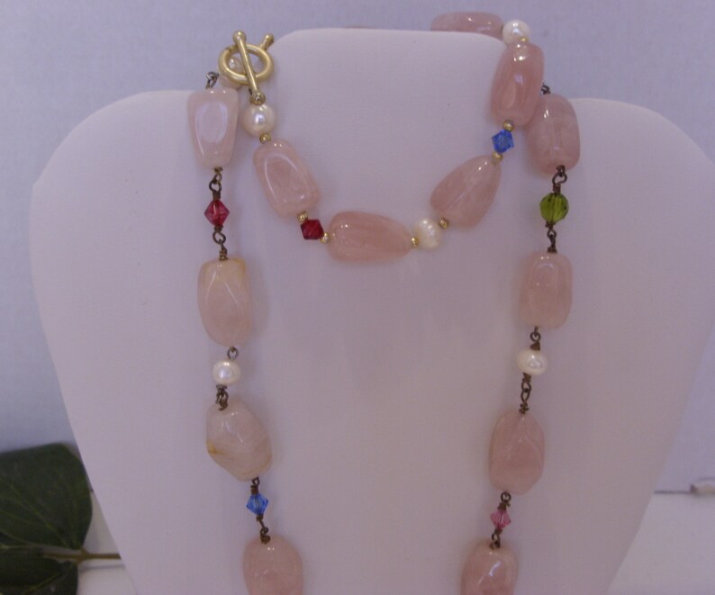 May include: A long necklace featuring large, light pink rose quartz beads, interspersed with small white pearls and colorful crystal accents. The necklace has a gold-tone toggle clasp.