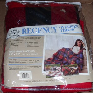 Op de afbeelding: Een rood, zwart en blauw geruite oversized plaid, 137 cm x 183 cm, in een doorzichtige plastic verpakking. De verpakking bevat het woord "REGENCY" en een foto van een vrouw die de plaid gebruikt. De plaid is gemaakt van 100% virgin acryl.