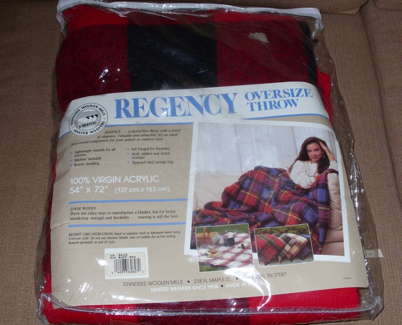Op de afbeelding: Een rood, zwart en blauw geruite oversized plaid, 137 cm x 183 cm, in een doorzichtige plastic verpakking. De verpakking bevat het woord "REGENCY" en een foto van een vrouw die de plaid gebruikt. De plaid is gemaakt van 100% virgin acryl.