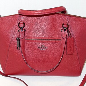 May include: A red leather handbag with a top handle and a detachable shoulder strap. The bag has a front zipper pocket and a metal logo. The bag has silver-tone hardware and a matching leather tag.