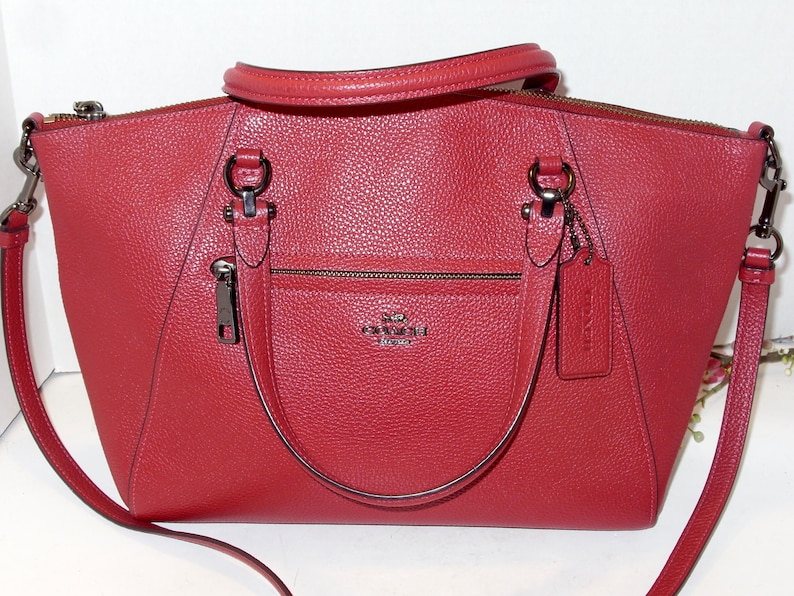May include: A red leather handbag with a top handle and a detachable shoulder strap. The bag has a front zipper pocket and a metal logo. The bag has silver-tone hardware and a matching leather tag.