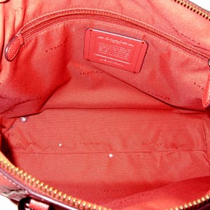 May include: Open view of a red leather handbag interior. The bag features a zippered top, a red fabric lining, and a small rectangular tag. The interior includes a zippered pocket and a large open space. The zipper is gold-colored.