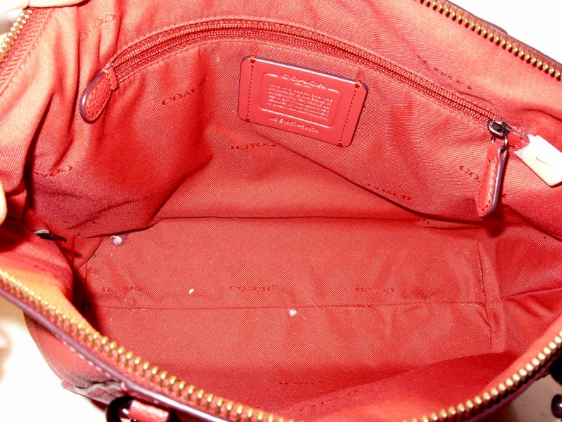 May include: Open view of a red leather handbag interior. The bag features a zippered top, a red fabric lining, and a small rectangular tag. The interior includes a zippered pocket and a large open space. The zipper is gold-colored.