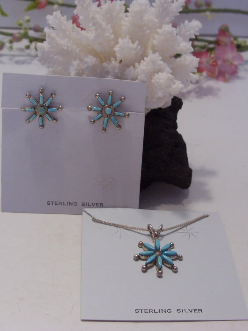 May include: A set of sterling silver earrings and a necklace featuring turquoise flower designs. The earrings and pendant are both in the shape of a starburst with turquoise stones set in the center and around the edges.