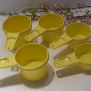 May include: A set of four bright yellow plastic measuring cups with round bowls and flat handles. Each handle features a small circular hole at the end for hanging. The cups vary in size and are arranged in a cluster, showcasing their smooth finish.