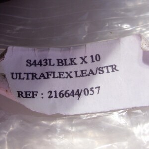 Puede incluir: Una etiqueta blanca rasgada con texto negro. La etiqueta dice "S443L BLK X 10 ULTRAFLX LEA/STR REF: 216644/057". La etiqueta est&aacute; parcialmente oscurecida por un material pl&aacute;stico transl&uacute;cido y arrugado.