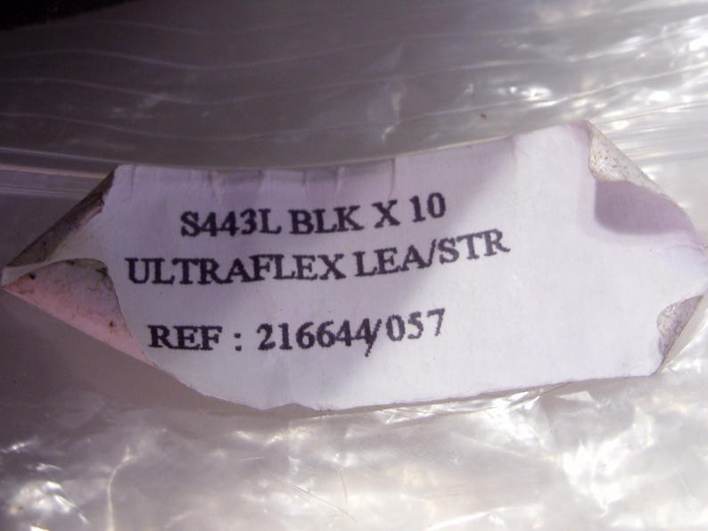 Puede incluir: Una etiqueta blanca rasgada con texto negro. La etiqueta dice "S443L BLK X 10 ULTRAFLX LEA/STR REF: 216644/057". La etiqueta est&aacute; parcialmente oscurecida por un material pl&aacute;stico transl&uacute;cido y arrugado.