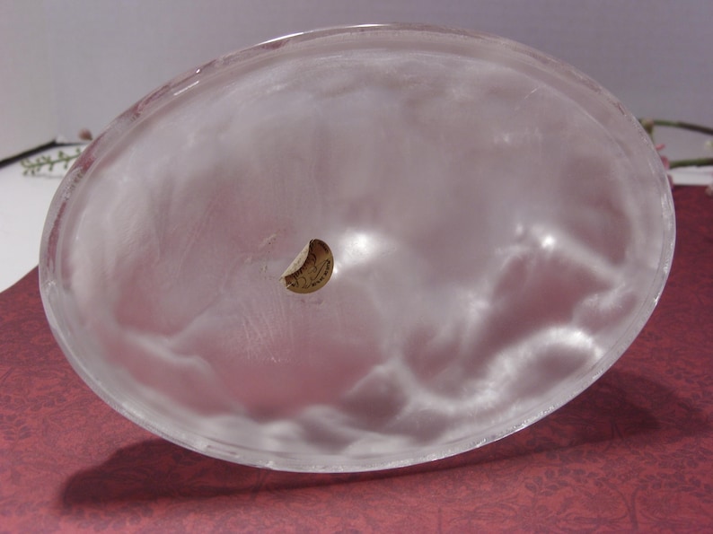 May include: A clear, oval-shaped glass bowl with a frosted appearance. The bowl has a small, circular sticker on the inside. The bowl is likely used for decorative purposes or serving food.