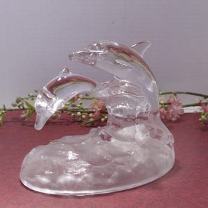 May include: A clear glass sculpture of two dolphins leaping from a wave. The dolphins are detailed with fins and facial features. The sculpture is set against a soft background with floral accents.