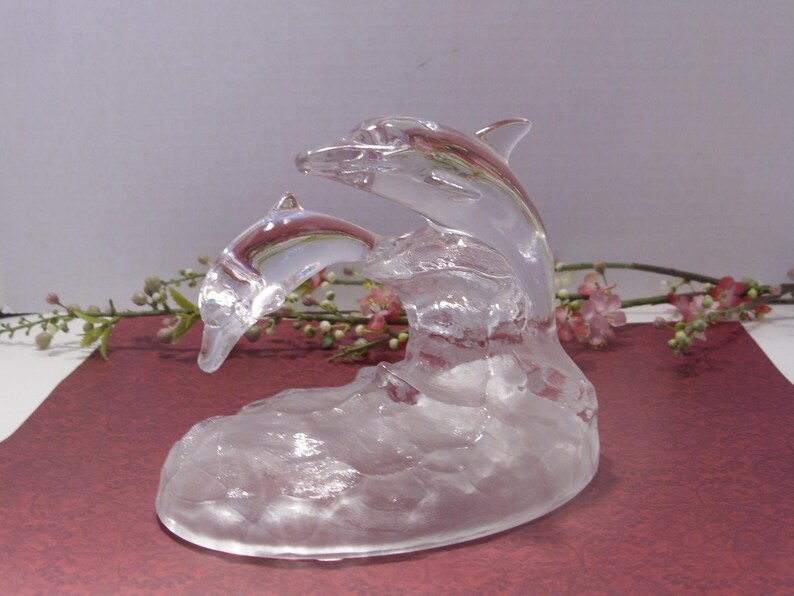 May include: A clear glass sculpture of two dolphins leaping from a wave. The dolphins are detailed with fins and facial features. The sculpture is set against a soft background with floral accents.