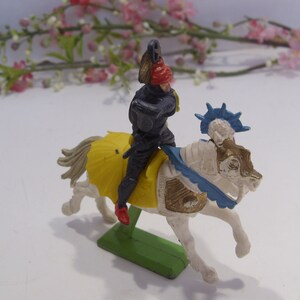 May include: A plastic toy knight riding a white horse with a blue and gold saddle. The knight is wearing black armor and a red helmet. The horse has a blue and white crest.