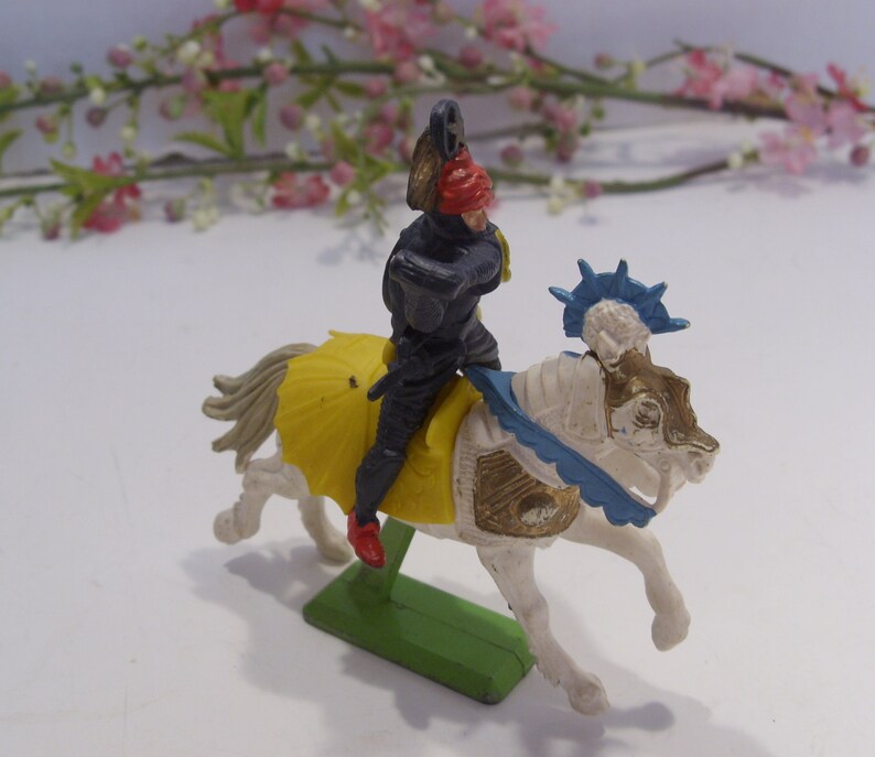 May include: A plastic toy knight riding a white horse with a blue and gold saddle. The knight is wearing black armor and a red helmet. The horse has a blue and white crest.