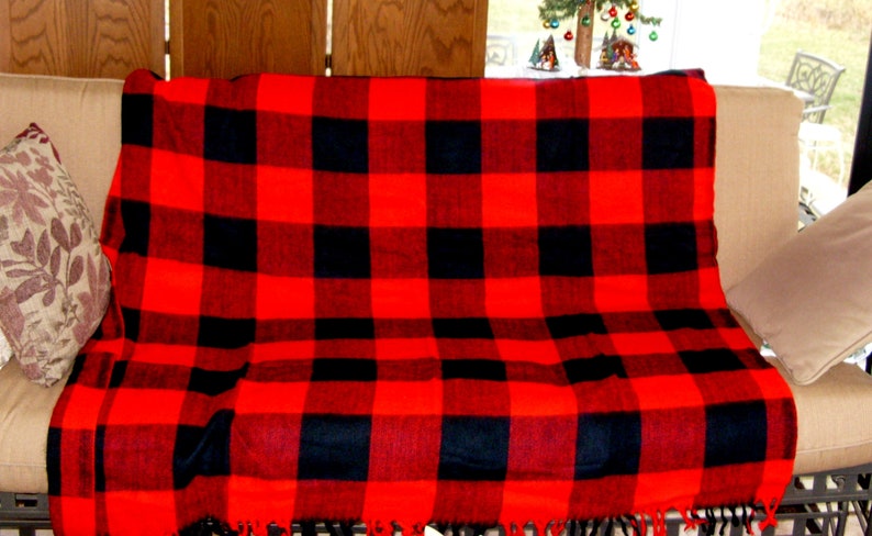 Op de afbeelding: Een rood-zwarte geruite deken, met franjes, gedrapeerd over een beige buitenzitbank. Twee decoratieve kussens zijn aan weerszijden van de deken geplaatst. Het patroon is een klassieke buffelruit.
