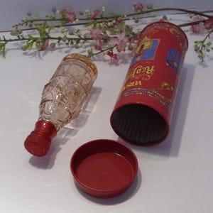 May include: A clear glass bottle with a red screw cap, a cylindrical red metal container with the text Versace Red Jeans, and a separate red metal lid. The bottle features a textured surface and is positioned next to the open container.