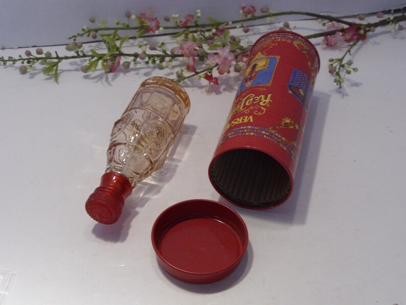 May include: A clear glass bottle with a red screw cap, a cylindrical red metal container with the text Versace Red Jeans, and a separate red metal lid. The bottle features a textured surface and is positioned next to the open container.