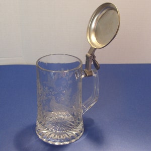 May include: Clear glass beer stein with etched pheasant and foliage design. Features a hinged metal lid and handle. The base has a decorative, scalloped edge. A classic drinking vessel.