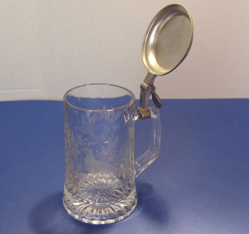 May include: Clear glass beer stein with etched pheasant and foliage design. Features a hinged metal lid and handle. The base has a decorative, scalloped edge. A classic drinking vessel.