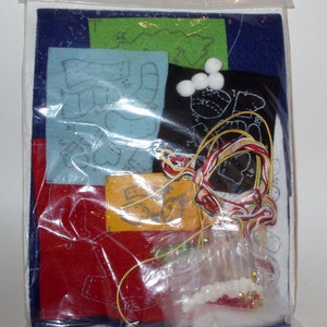 May include: A craft kit containing felt pieces in various colors, including blue, red, and green, with printed patterns. The kit also includes white pom-poms, gold and multi-colored thread, and small beads. The kit is packaged in a clear plastic bag.