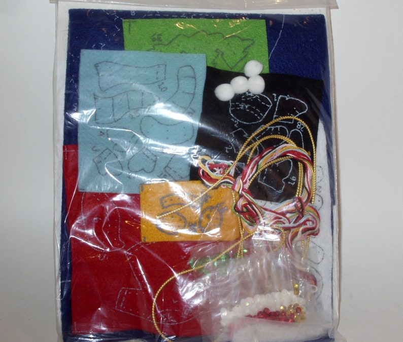 May include: A craft kit containing felt pieces in various colors, including blue, red, and green, with printed patterns. The kit also includes white pom-poms, gold and multi-colored thread, and small beads. The kit is packaged in a clear plastic bag.