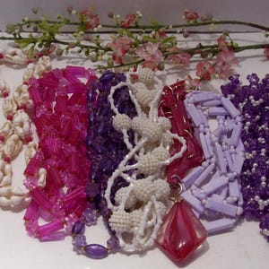 May include: A collection of six necklaces featuring various designs: a white seashell strand, a bright pink tubular bead necklace, a deep purple faceted bead strand, a white textured ball bead necklace, a light lavender rectangular bead strand, and a dark purple beaded necklace with a large red diamond-shaped pendant.