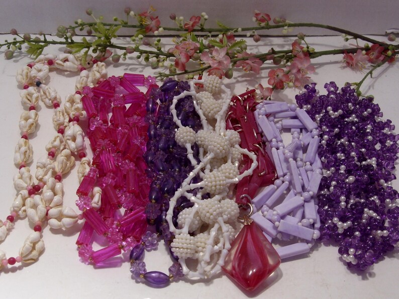 May include: A collection of six necklaces featuring various designs: a white seashell strand, a bright pink tubular bead necklace, a deep purple faceted bead strand, a white textured ball bead necklace, a light lavender rectangular bead strand, and a dark purple beaded necklace with a large red diamond-shaped pendant.