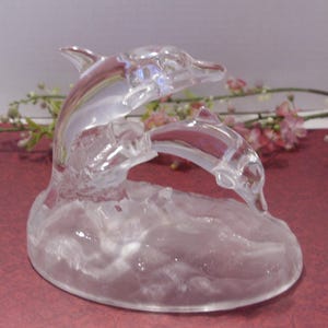 May include: A clear glass sculpture of two dolphins leaping over waves. The dolphins are detailed with fins and a curved body, and the base has a textured wave design. The sculpture is set against a background of flowers.