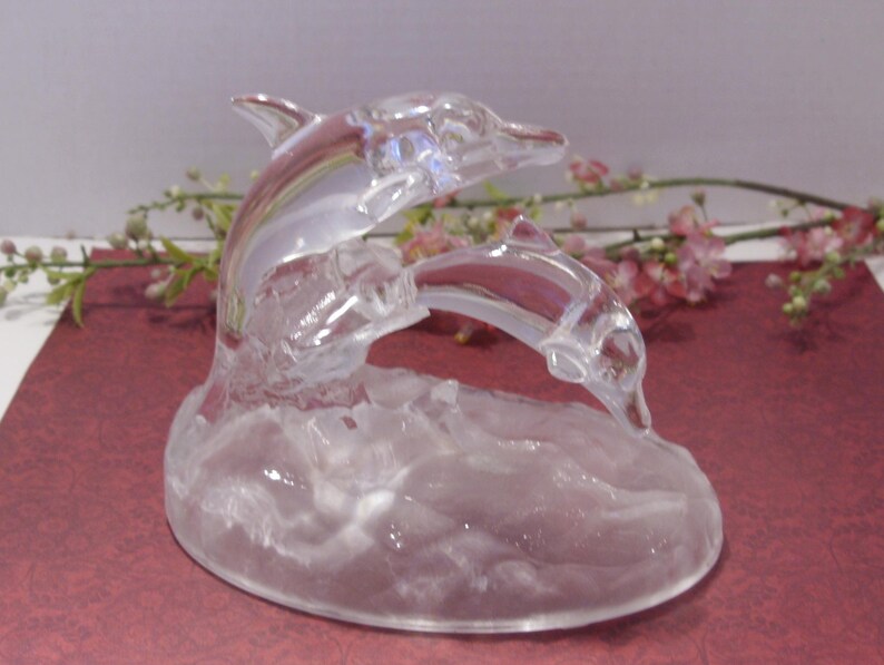 May include: A clear glass sculpture of two dolphins leaping over waves. The dolphins are detailed with fins and a curved body, and the base has a textured wave design. The sculpture is set against a background of flowers.