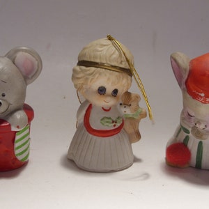 May include: Three ceramic Christmas ornaments. The ornaments include a gray mouse in a red pot, a girl holding a teddy bear, and a mouse wearing a red hat and striped pajamas. Each ornament has a gold string for hanging.
