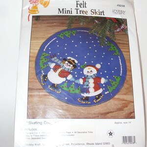May include: A felt mini tree skirt kit with a blue background featuring two snow people skating. The design includes green trees and white snowflakes. The package reads "Felt Mini Tree Skirt" and is approximately 14 inches in diameter.