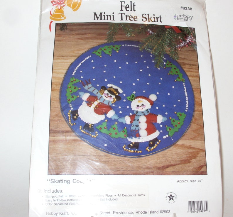 May include: A felt mini tree skirt kit with a blue background featuring two snow people skating. The design includes green trees and white snowflakes. The package reads "Felt Mini Tree Skirt" and is approximately 14 inches in diameter.