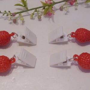 Op de afbeelding: Vier witte plastic clips met rode aardbei bedels. De bedels zijn getextureerd en bevestigd aan de clips met kleine zilverkleurige ringen. Een takje bloemen staat op de achtergrond.
