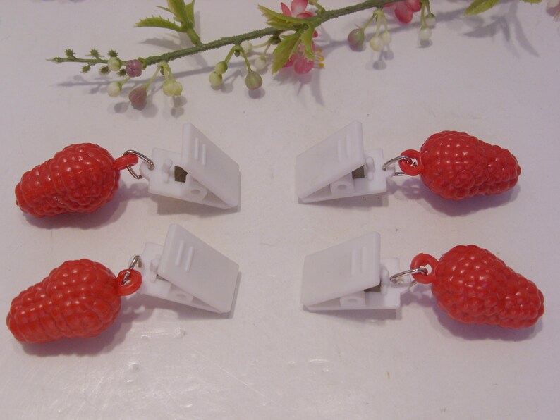 Op de afbeelding: Vier witte plastic clips met rode aardbei bedels. De bedels zijn getextureerd en bevestigd aan de clips met kleine zilverkleurige ringen. Een takje bloemen staat op de achtergrond.