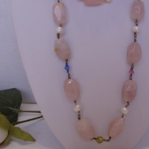 May include: A necklace and bracelet set featuring large, light pink, oval-shaped beads. The necklace has small white pearls and colorful accents. The jewelry is displayed on a white mannequin, with a rose and greenery in the foreground.