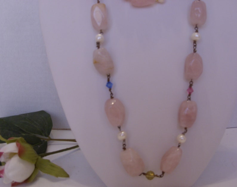 May include: A necklace and bracelet set featuring large, light pink, oval-shaped beads. The necklace has small white pearls and colorful accents. The jewelry is displayed on a white mannequin, with a rose and greenery in the foreground.