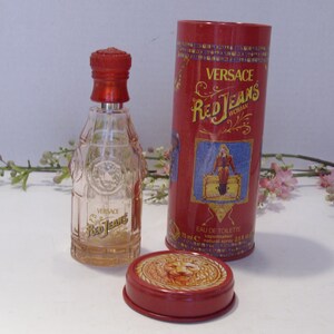 May include: A clear glass Versace Red Jeans eau de toilette bottle with a red cap, accompanied by a matching red cylindrical metal canister and a separate red lid featuring a lion emblem. The canister displays the text Versace Red Jeans Woman.