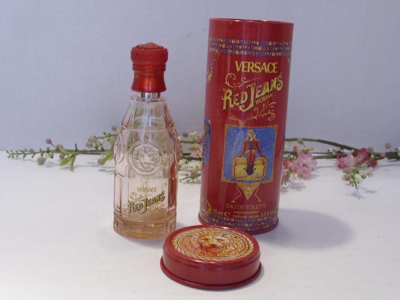 May include: A clear glass Versace Red Jeans eau de toilette bottle with a red cap, accompanied by a matching red cylindrical metal canister and a separate red lid featuring a lion emblem. The canister displays the text Versace Red Jeans Woman.