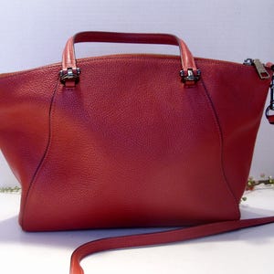May include: A red leather handbag with a top handle and a detachable shoulder strap. The bag has a zipper closure and silver-tone hardware. The leather has a textured appearance. The bag is a medium size.