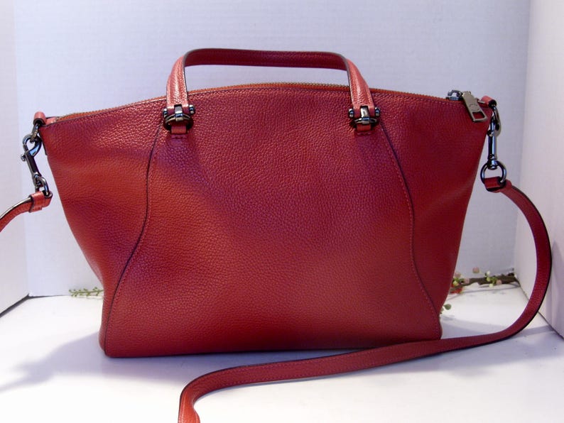 May include: A red leather handbag with a top handle and a detachable shoulder strap. The bag has a zipper closure and silver-tone hardware. The leather has a textured appearance. The bag is a medium size.