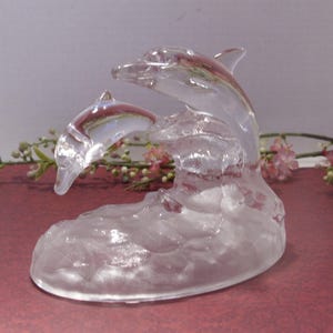 May include: A clear glass sculpture of two dolphins leaping from a wave. The dolphins are detailed with fins and curved bodies, and the wave base has a textured appearance. The sculpture is set against a soft background.
