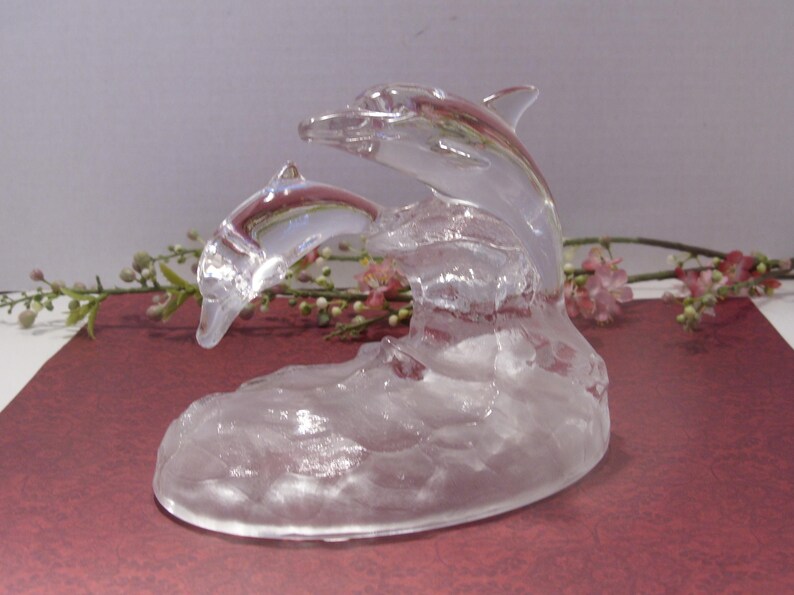 May include: A clear glass sculpture of two dolphins leaping from a wave. The dolphins are detailed with fins and curved bodies, and the wave base has a textured appearance. The sculpture is set against a soft background.