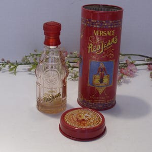 May include: A clear glass perfume bottle with a red cap and the text Versace Red Jeans, accompanied by a matching red cylindrical metal tin featuring the text Versace Red Jeans Woman, an illustration of a figure, and a lion emblem on the lid.