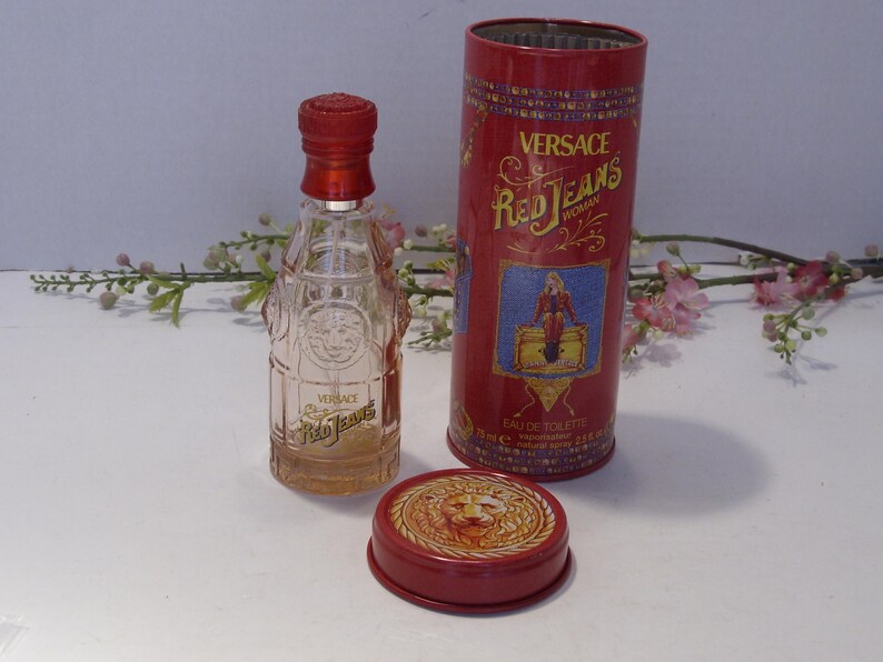 May include: A clear glass perfume bottle with a red cap and the text Versace Red Jeans, accompanied by a matching red cylindrical metal tin featuring the text Versace Red Jeans Woman, an illustration of a figure, and a lion emblem on the lid.