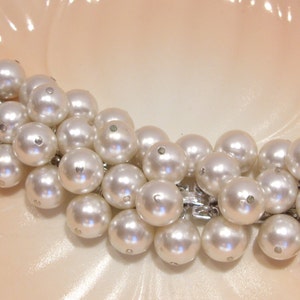 May include: A vintage pearl bracelet featuring multiple strands of large, round, lustrous white pearls. The pearls are connected by silver-toned metal links. The bracelet is displayed on a light peach-colored surface.
