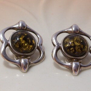May include: A pair of silver earrings with a green amber stone in the center of each earring. The earrings are shaped like a flower with four petals.
