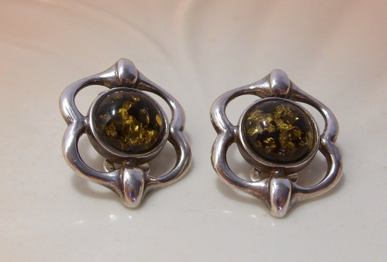May include: A pair of silver earrings with a green amber stone in the center of each earring. The earrings are shaped like a flower with four petals.