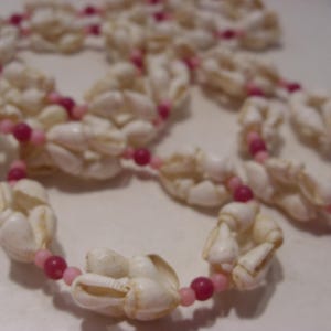 May include: A long necklace constructed from white sea shells interspersed with small, round beads in alternating shades of light pink and dark magenta. The shells are arranged in clusters along the length of the strand, creating a textured pattern.