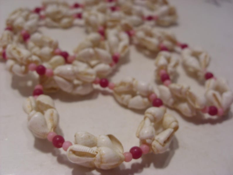 May include: A long necklace constructed from white sea shells interspersed with small, round beads in alternating shades of light pink and dark magenta. The shells are arranged in clusters along the length of the strand, creating a textured pattern.