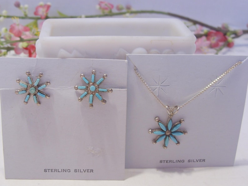 May include: A set of sterling silver earrings and a necklace with turquoise flower pendants. The earrings and pendant are both in the shape of a flower with a turquoise center and silver petals. The cards say "Sterling Silver".